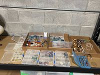 Photo showing several cardboard boxes and plastic organizer boxes filled with assorted costume jewelry including necklaces, earrings, and jewelry watches.