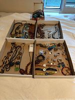 Top view of costume jewelry assortment in four cardboard trays including necklaces, earrings, bracelets, watches, and brooches.