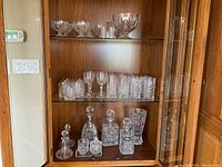 Wide cabinet shelf view of multiple cut crystal glasses, stemware, decanters with stoppers, and a crystal vase on shelves.