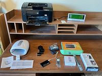 Overview shot of all lot items on wooden desk including HP printer, clock radio, massager, cameras, watches, flashlight, and boxed phone.
