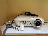 White Epson projector with lens on right, mounting bracket on left, and black power cord coiled on top, sitting on white surface.