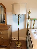Full view of brass floor lamp with beige fabric shade next to wooden dresser and brass bed frame, showing overall height and design.
