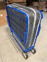 Folding cot folded with mattress, showing blue metal frame and mattress thickness and pattern.