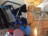 Photo showing blue rollator walker with seat and basket, silver standard walker, and canes grouped against a wall alongside cushions and bed assist rail.