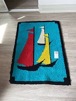 Full view of hooked mat with three sailboats in red, yellow, and white sails on a turquoise background with black border, placed on wood floor.