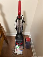 Front view of Hoover PowerDash upright carpet cleaner with red handle, cord wrapped around the handle, clear water tanks next to it, user manual and clear cleaning accessory on the floor.