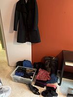 Full view of various clothing items including scarves, sweaters, two pairs of slippers, shoes, and travel bags on the floor and hanging black coat on the wall.