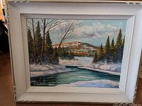Full front image of one oil painting featuring a winter landscape with snowy banks and a river under a cloudy sky, framed in an ornate white frame with visible wear on corners.