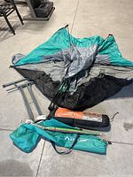 Collapsed teal, gray, and black sun shelter with collapsible frame components and storage bags on concrete floor.