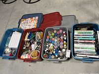Four plastic storage bins side by side, containing art materials such as paint bottles, canvases, brushes, and completed artworks.