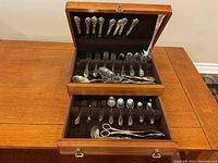 Open wooden canteen displaying all flatware pieces arranged in their slots on two tiers, silver plated with floral Noritake Serenade pattern handles.