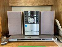 Full shot of Sony CMT-EH10 system with two speakers placed on a wooden surface alongside a silver LG DVD player and two remote controls.