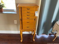 Closed honey oak veneer standing jewelry case with five front drawers and side doors closed, showing overall front view with metal handles and curved legs.