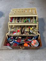 Open fishing tackle box with trays extended showing various fishing lures, hooks, floats, fishing line, and accessories inside compartments