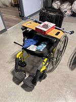 Manual Invacare wheelchair with wooden tray, heating bags, box of Sunbeam cordless heating pad, and cane placed across the backrest.