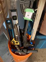 Orange bucket containing various tools including two spirit levels (one yellow Stanley), several C-clamps with blue handles, a large metal framing square, and a handsaw with a wooden handle.