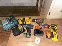Photo showing an assortment of hand tools including drills, chargers, batteries, and laser measuring devices on a wooden floor.