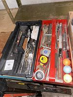 Photo showing a black plastic tool tray with assorted hand tools such as drill bits, screwdriver, wrench, and a metal extension bar. Adjacent red tray contains clamps, tape measures, and golf accessories including golf balls and tees.