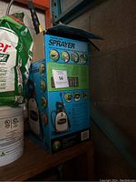 Photo showing blue and white premium sprayer box with images and instructions on the side, placed on a shelf next to garden supplies.