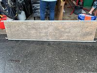 Photo of a single wooden scaffold board lying horizontally on asphalt with metal support bar underneath and people in the background.