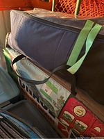 A dark blue fabric storage bag for camping gear, resting on boxes and plastic containers, secured with green tape and black straps.