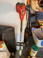 Full view of the red and black Honda weed whacker with an orange top, leaning against a wall with associated workbench items in background.