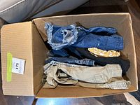 Box containing multiple women's pants, capris, and shorts in assorted colors and styles, showing denim, tan, black, and patterned fabrics.