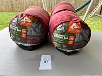 Two rolled and compressed maroon sleeping bags with gray labels and black carrying sacks facing forward showing brand and specifications.