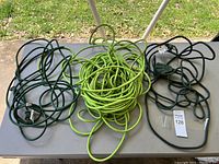 Three extension cords arranged on a table outdoors, showing two green and one black cord.