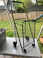 Collapsible wire mesh utility cart shown from the side with handle and wheels on a table.