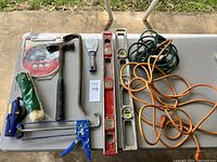 Top view of assorted hand tools including levels, hammer, caulking gun, and extension cords arranged on a white table outdoors.