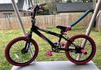 Side view of black and pink kids bike with pink wheels and pedals, and padded seat with blue and pink accents. Bike is standing on a concrete porch with a grassy yard and wooden fence in background.
