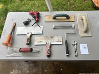 View of the lot on a grey table showing various plastering and drywall tools, including trowels, cordless drill, wrench, and paint roller frame.