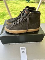 Side view of pair of dark-colored Nike high-top sneakers on black shoebox outdoors, showing gum sole, laces, and Nike swoosh on side.