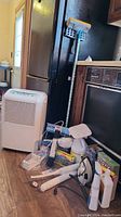 Photo showing dehumidifier, mop, cleaning supplies, steamer and iron arranged on floor near kitchen area.
