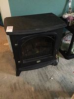 Front view of black Dimplex electric fireplace stove with a glass door and brass handles, positioned on carpeted floor.