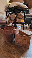 View of rattan harvest basket and wooden Canadian butter box together on floor, basket is tall with handles, wooden box rectangular with text on side.