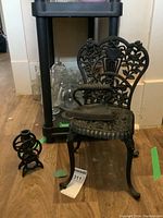 Small decorative iron chair with intricate floral and heart patterns in backrest and seat, black finish, shown on wood floor with glassware in background.