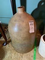 Full view of large beige stoneware jug standing on floor, showing rounded body, narrow neck, and chipped spout.