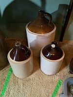 Three ceramic jugs arranged on carpet; varying sizes; chips and cracks visible; brown glazed tops and white bodies.