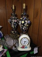 Pair of porcelain table lamps with black and floral pattern and vintage porcelain clock with floral decoration on wooden surface.