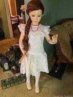 Full view of child size mannequin in white lace dress, tights, cream shoes, and necklace with braided wig.