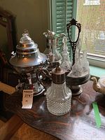 Full view of the set showing electric coffee pot, coffee carafe, condiment server with multiple bottles, and silverplate topped glass pitcher on wooden table near window.