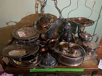 Photo showing a variety of tarnished silverplate serveware items, including trays, pots, and bowls arranged on a wooden table.