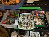 Tray with multiple small Wade ceramic figurines, salt and pepper shakers, and other small figurines in plastic containers on a shelving unit.