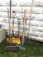 Photo showing a variety of yard tools arranged against a white brick wall on grass, including hand cultivators, rakes, a broom, a shovel, and a snow shovel.