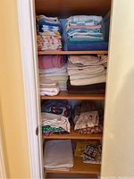 Closet with organized shelves holding bath towels, hand towels, face cloths, aprons, tablecloths, and decorative fabric items.