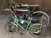 Three vintage cruiser bicycles grouped together in barn setting. Green Mustang bike visible in front with black grips and pedals, blue Vagabond in middle, burgundy New Hudson in back. Bikes show wear and surface rust.