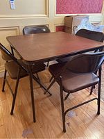 Brown metal folding card table set up with four matching chairs around it on hardwood floor.