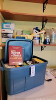 Photo showing a blue Rubbermaid tote filled with cleaning supplies and The Queen of Clean's cleaning guide book on top, with shelves of additional cleaning products in the background.